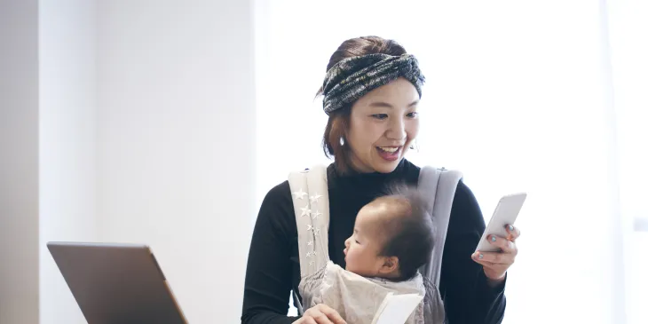 Woman with baby in carrier, using mobile phone and working